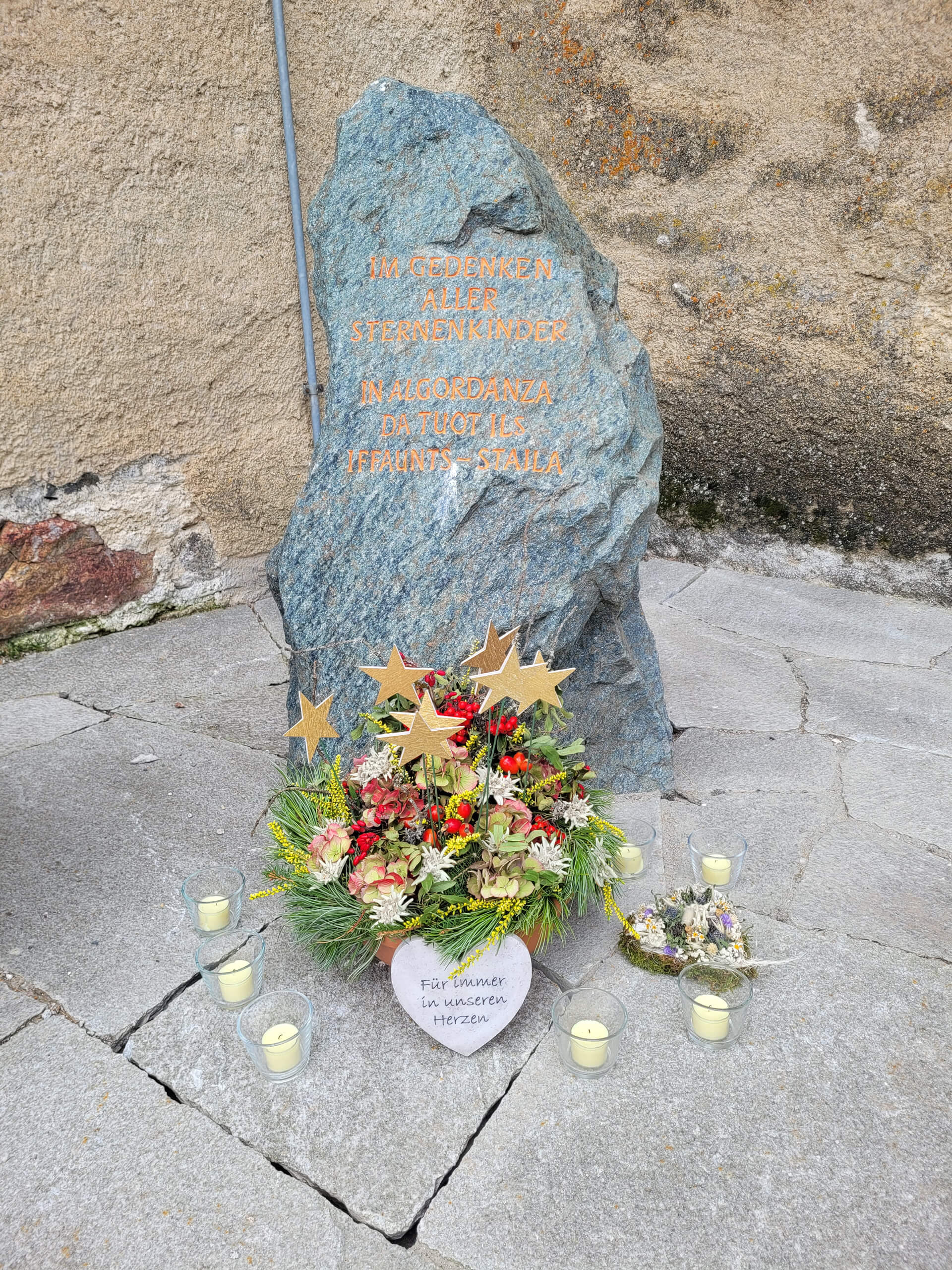 Sternenkindgrab in Celerina mit einem Gedenkstein, Blumenkranz und Kerzen